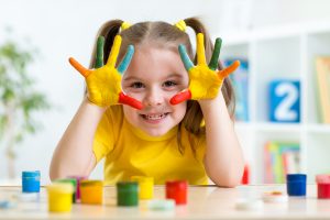 little girl in yellow shirt with brightly painted hands smiling at camera